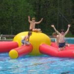 Vier Kinder spielen auf einer großen aufblasbaren Struktur in einem Schwimmbecken, wobei zwei Kinder abspringen und zwei andere auf die aufblasbare Struktur klettern. Im Hintergrund sind Bäume zu sehen.