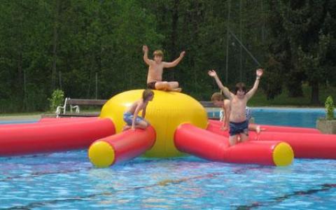 Vier Kinder spielen auf einer großen aufblasbaren Struktur in einem Schwimmbecken, wobei zwei Kinder abspringen und zwei andere auf die aufblasbare Struktur klettern. Im Hintergrund sind Bäume zu sehen.