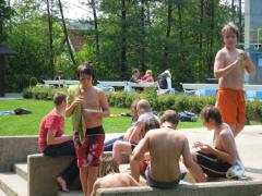 Mehrere hemdsärmelige Jungen versammeln sich im Freien in der Nähe eines Pools, einige sitzen auf Betonstufen, andere stehen, mit Bäumen und weiteren Menschen im Hintergrund.