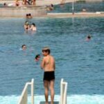 Eine Person steht auf einem Sprungbrett über einem Schwimmbad, während im Hintergrund mehrere Personen im Wasser schwimmen und sich auf den Beckenrändern entspannen.