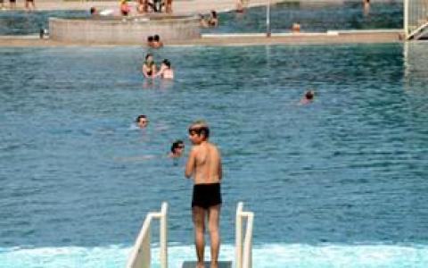 Eine Person steht auf einem Sprungbrett über einem Schwimmbad, während im Hintergrund mehrere Personen im Wasser schwimmen und sich auf den Beckenrändern entspannen.