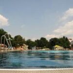 Freibad mit Menschen, die sich entspannen und schwimmen, umgeben von Bäumen und Gebäuden unter einem teilweise bewölkten Himmel.