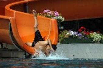 Eine Person in schwarzer Badehose verlässt kopfüber eine orangefarbene Wasserrutsche und spritzt in ein Becken; im Hintergrund sind Blumen in Pflanzgefäßen zu sehen.