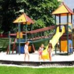 Drei Kinder spielen auf einem Spielplatz mit einer gelben Rutsche und Klettergerüsten, umgeben von Gras und Bäumen an einem sonnigen Tag.