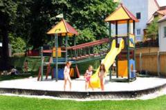 Drei Kinder spielen auf einem Spielplatz mit einer gelben Rutsche und Klettergerüsten, umgeben von Gras und Bäumen an einem sonnigen Tag.