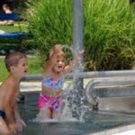 Zwei kleine Kinder in Badeanzügen spielen und planschen an einem sonnigen Tag am Rande eines flachen Freibads.