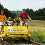 Eine vierköpfige Familie fährt mit einem gelben Schienenfahrrad auf Bahngleisen durch eine ländliche Landschaft mit Gras und Bäumen.