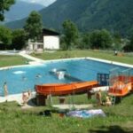 Freibad mit schwimmenden und spielenden Menschen, umgeben von Gras, Bäumen und Bergen; im Vordergrund sind zwei orangefarbene Wasserrutschen zu sehen.