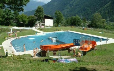 Freibad mit schwimmenden und spielenden Menschen, umgeben von Gras, Bäumen und Bergen; im Vordergrund sind zwei orangefarbene Wasserrutschen zu sehen.