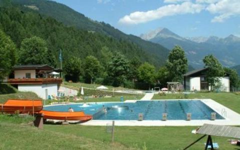 Freibad, umgeben von Gras, Bäumen und Bergen unter einem teilweise bewölkten Himmel, mit mehreren Gebäuden in der Nähe und einer Rutsche, die in den Pool führt.