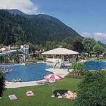 Ein von Gras und Bäumen umgebener Freibadkomplex mit sonnenbadenden Menschen, Bergen im Hintergrund und einem teilweise bewölkten Himmel.