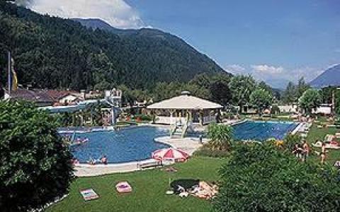 Ein von Gras und Bäumen umgebener Freibadkomplex mit sonnenbadenden Menschen, Bergen im Hintergrund und einem teilweise bewölkten Himmel.