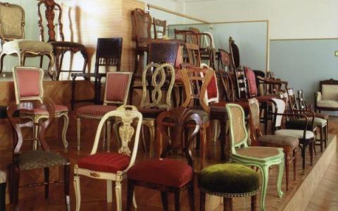 Eine Ausstellung verschiedener antiker Holzstühle mit gepolsterten Sitzen, die in Reihen auf gestuften Holzplattformen in einem Museum oder einer Galerie angeordnet sind.