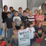 Eine Gruppe von Kindern und zwei Erwachsene posieren zusammen in einer Bibliothek. Einige Kinder halten ein großes Plakat mit der Aufschrift "Welt Woche".