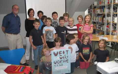 Eine Gruppe von Kindern und zwei Erwachsene posieren zusammen in einer Bibliothek. Einige Kinder halten ein großes Plakat mit der Aufschrift "Welt Woche".