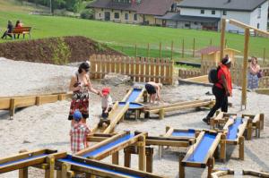 Mehrere Kinder und Erwachsene spielen mit einer Wasserspielanlage auf einem Spielplatz im Freien, mit Wiesen und Häusern im Hintergrund.