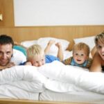 Ein Mann, eine Frau und zwei kleine Kinder liegen auf dem Bauch auf einem Bett in einem hell erleuchteten Raum und lächeln in die Kamera.
