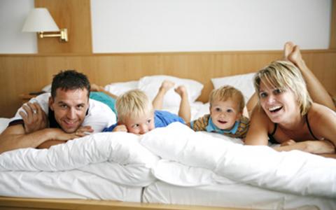 Ein Mann, eine Frau und zwei kleine Kinder liegen auf dem Bauch auf einem Bett in einem hell erleuchteten Raum und lächeln in die Kamera.