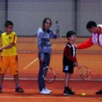 Ein Tennistrainer gibt drei Kindern, die auf einem Tennisplatz stehen und jeweils einen Schläger halten, Anweisungen.