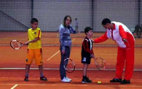 Ein Tennistrainer gibt drei Kindern, die auf einem Tennisplatz stehen und jeweils einen Schläger halten, Anweisungen.