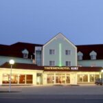Ein großes Hotelgebäude mit der beleuchteten Aufschrift "Thermenhotel Kurz", beleuchtet in der Abenddämmerung, mit parkenden Autos davor und einem klaren Abendhimmel.