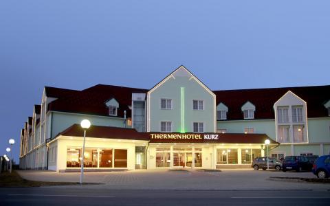 Ein großes Hotelgebäude mit der beleuchteten Aufschrift "Thermenhotel Kurz", beleuchtet in der Abenddämmerung, mit parkenden Autos davor und einem klaren Abendhimmel.