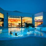 Freibad mit mehreren Personen, die in der Abenddämmerung schwimmen, mit einem hell erleuchteten modernen Gebäude mit großen Fenstern im Hintergrund.