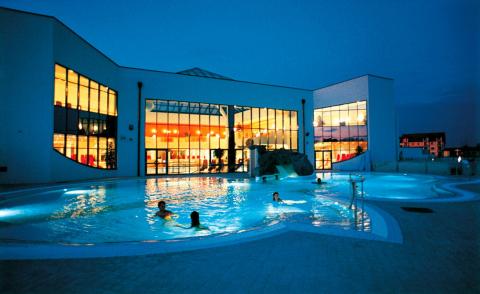 Freibad mit mehreren Personen, die in der Abenddämmerung schwimmen, mit einem hell erleuchteten modernen Gebäude mit großen Fenstern im Hintergrund.
