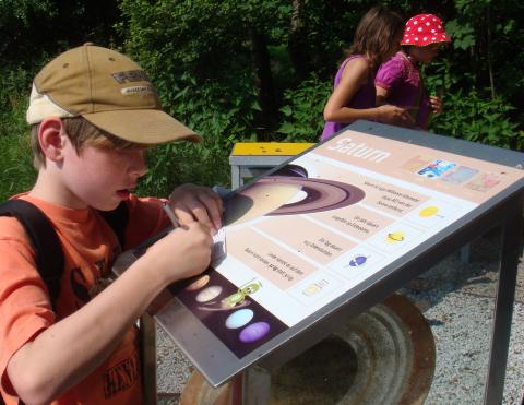 Ein Junge mit hellbrauner Mütze interagiert mit einem Saturn-Informationsdisplay im Freien; im Hintergrund stehen zwei Mädchen, von denen eines eine rote Tupfenmütze trägt.