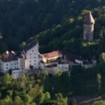 Luftaufnahme einer mittelalterlichen Burganlage mit Umfassungsmauern und einem hohen Rundturm, eingebettet in eine dicht bewaldete, hügelige Landschaft.