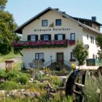 Ein weißes, zweistöckiges Haus mit der Aufschrift "Hildegard Naturhaus" mit Blumenkästen, umgeben von einem Garten und Grünanlagen, mit einem hölzernen Wasserrad im Vordergrund.
