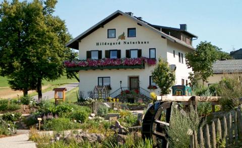 Ein weißes, zweistöckiges Haus mit der Aufschrift "Hildegard Naturhaus" mit Blumenkästen, umgeben von einem Garten und Grünanlagen, mit einem hölzernen Wasserrad im Vordergrund.