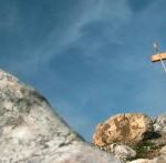 Ein Holzkreuz steht auf einer felsigen Anhöhe unter einem blauen Himmel mit Wolkenfetzen.