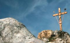 Ein Holzkreuz steht auf einer felsigen Anhöhe unter einem blauen Himmel mit Wolkenfetzen.