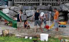 Fünf Kinder spielen zusammen in einem Sandgebiet mit Spielzeug und einer Rutsche, mit großen Felsen und einem Zaun im Hintergrund.