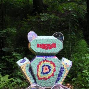 Eine farbenfrohe Mosaikskulptur eines Bären steht im Freien in einer üppigen, grünen Waldumgebung.