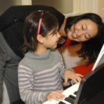 Ein junges Mädchen spielt Klavier, während ein Erwachsener sich vorbeugt und lächelt, möglicherweise um Anweisungen zu geben oder zu ermutigen. Auf dem Klavier sind Notenblätter zu sehen.