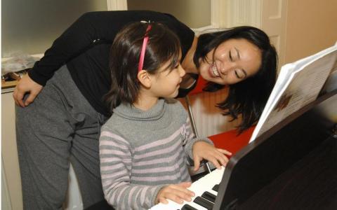 Ein junges Mädchen spielt Klavier, während ein Erwachsener sich vorbeugt und lächelt, möglicherweise um Anweisungen zu geben oder zu ermutigen. Auf dem Klavier sind Notenblätter zu sehen.