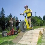 Ein Kind, das einen Helm trägt, fährt mit einem gelben Roller über eine Holzrampe im Freien, mit Menschen und Bäumen im Hintergrund.