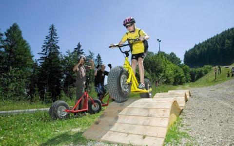Ein Kind, das einen Helm trägt, fährt mit einem gelben Roller über eine Holzrampe im Freien, mit Menschen und Bäumen im Hintergrund.