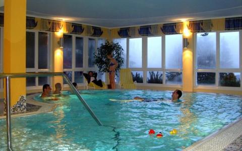 Mehrere Personen schwimmen und entspannen sich in einem Hallenbad mit großen Fenstern und gelben Wänden; Poolspielzeug schwimmt auf dem Wasser.