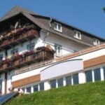 Ein dreistöckiges Gebäude mit Blumenkästen auf hölzernen Balkonen, weißen Wänden, großen Fenstern und einem schrägen Dach, auf einem grasbewachsenen Hügel unter einem klaren blauen Himmel.