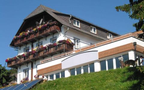 Ein dreistöckiges Gebäude mit Blumenkästen auf hölzernen Balkonen, weißen Wänden, großen Fenstern und einem schrägen Dach, auf einem grasbewachsenen Hügel unter einem klaren blauen Himmel.