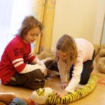 Zwei Kinder sitzen auf einem Holzboden und spielen mit einem langen Plüschtiger. Andere Plüschtiere und eine Puppe sind um sie herum in einem hell erleuchteten Raum verstreut.
