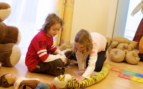 Zwei Kinder sitzen auf einem Holzboden und spielen mit einem langen Plüschtiger. Andere Plüschtiere und eine Puppe sind um sie herum in einem hell erleuchteten Raum verstreut.
