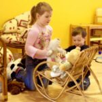 Zwei Kinder spielen mit Plüschtieren, die sie auf einen kleinen hölzernen Schaukelstuhl in einem hell erleuchteten Raum mit gelben Wänden und Holzmöbeln setzen.