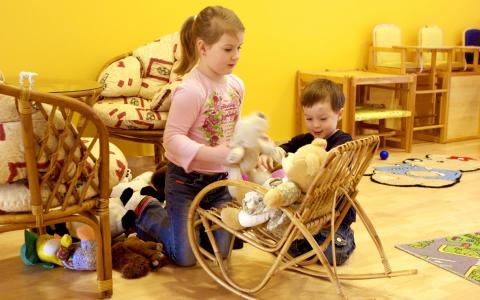 Zwei Kinder spielen mit Plüschtieren, die sie auf einen kleinen hölzernen Schaukelstuhl in einem hell erleuchteten Raum mit gelben Wänden und Holzmöbeln setzen.