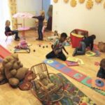 Mehrere kleine Kinder spielen mit Spielzeug, Puzzles und einer Rutsche in einem hell erleuchteten Spielzimmer, das mit Sonnenmotiven dekoriert und mit Plüschtieren und kleinen Möbeln ausgestattet ist.