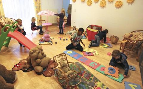 Mehrere kleine Kinder spielen mit Spielzeug, Puzzles und einer Rutsche in einem hell erleuchteten Spielzimmer, das mit Sonnenmotiven dekoriert und mit Plüschtieren und kleinen Möbeln ausgestattet ist.