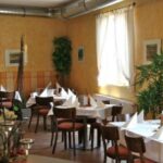 Ein kleiner Restaurantbereich mit mehreren Tischen, die mit weißen Tischdecken und Servietten gedeckt sind, Holzstühlen, Wandkunst und Topfpflanzen an den sonnenbeschienenen Fenstern.